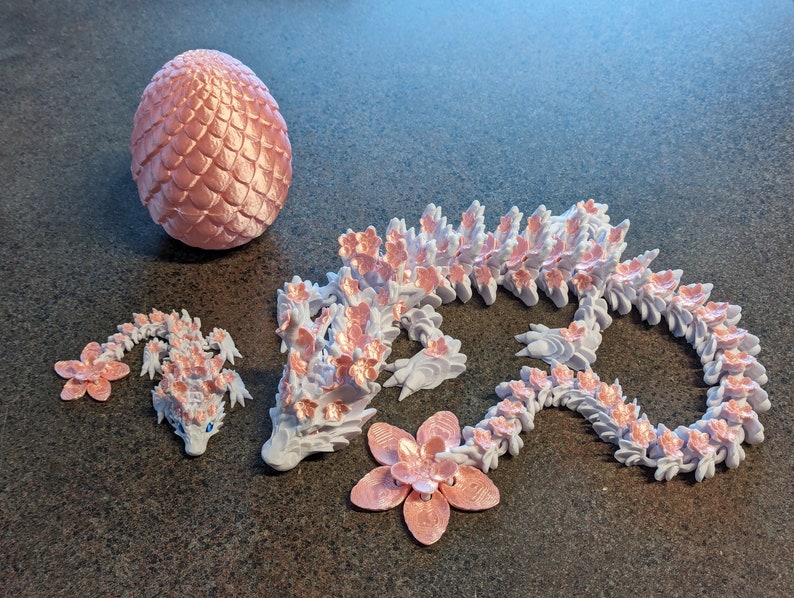 Puede incluir: Un conjunto de tres figuras de drag&oacute;n impresas en 3D de color rosa y blanco. El drag&oacute;n m&aacute;s grande est&aacute; enroscado en un c&iacute;rculo, el drag&oacute;n mediano est&aacute; tumbado y el drag&oacute;n m&aacute;s peque&ntilde;o est&aacute; de pie sobre sus patas traseras. Los dragones est&aacute;n todos decorados con flores rosas.