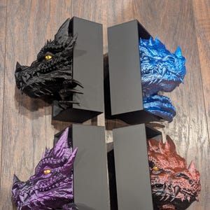 May include: Four dragon head bookends in black, blue, purple, and red. Each dragon head is emerging from a black rectangular box. The dragon heads have detailed scales and yellow eyes. The bookends are displayed on a wooden surface.