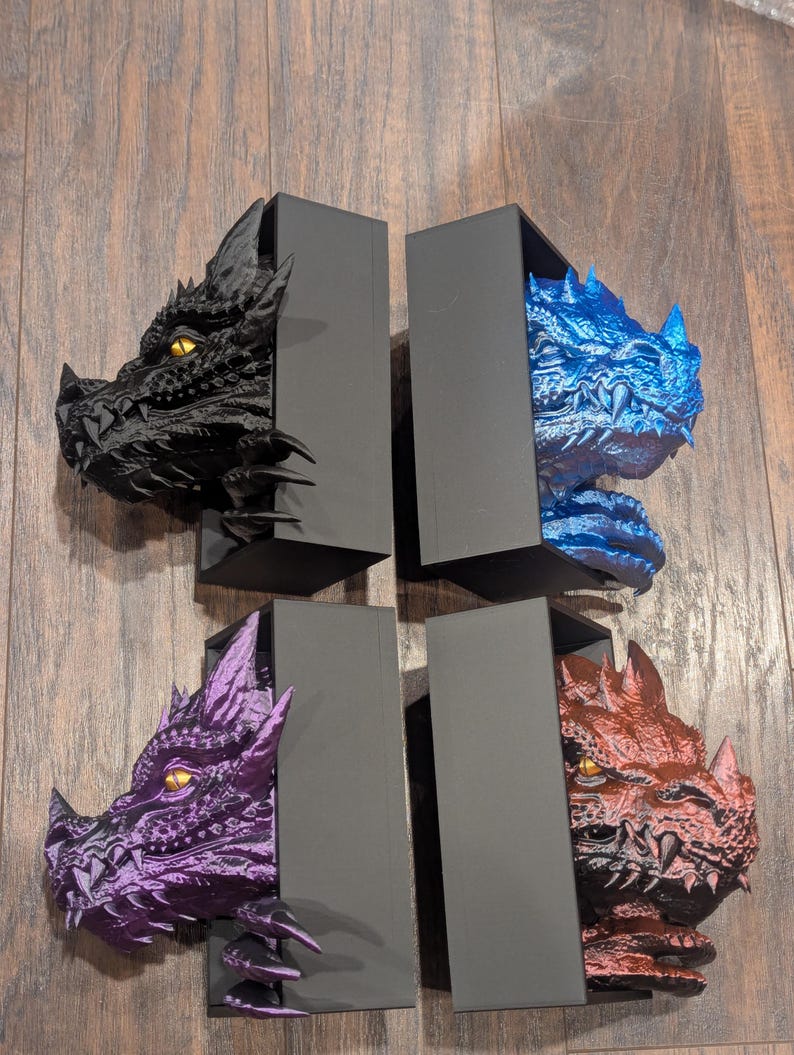 May include: Four dragon head bookends in black, blue, purple, and red. Each dragon head is emerging from a black rectangular box. The dragon heads have detailed scales and yellow eyes. The bookends are displayed on a wooden surface.
