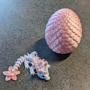 Puede incluir: Un huevo de drag&oacute;n rosa y una peque&ntilde;a figura de drag&oacute;n rosa y blanca. El huevo de drag&oacute;n est&aacute; hecho de un material texturizado que se asemeja a escamas. La figura de drag&oacute;n tiene una cola con forma de flor.