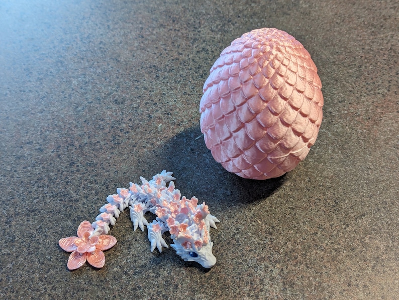 Puede incluir: Un huevo de drag&oacute;n rosa y una peque&ntilde;a figura de drag&oacute;n rosa y blanca. El huevo de drag&oacute;n est&aacute; hecho de un material texturizado que se asemeja a escamas. La figura de drag&oacute;n tiene una cola con forma de flor.