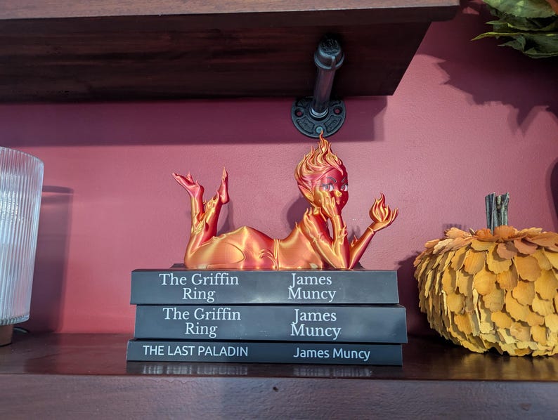 May include: A 3D-printed figurine in shades of orange and red, posed atop a stack of books. The figurine is of a person with a flame-like head, holding a small flame. The books are titled "The Griffin Ring" and "The Last Paladin".