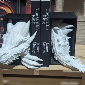 May include: White dragon head bookends. The bookends are made of a textured material and feature detailed dragon heads with sharp teeth and horns. The bookends are holding two black books with the title "The Griffin Ring" and the author's name "James Muncy".