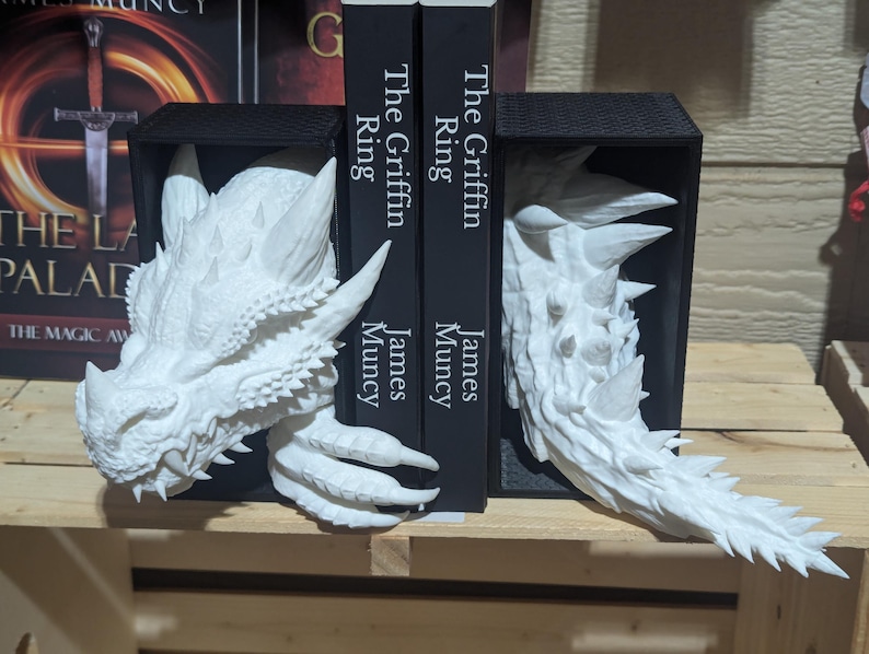 May include: White dragon head bookends. The bookends are made of a textured material and feature detailed dragon heads with sharp teeth and horns. The bookends are holding two black books with the title "The Griffin Ring" and the author's name "James Muncy".
