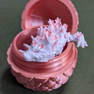 May include: A white, 3D printed dragon figurine with pink flowers on its back, inside a pink, 3D printed acorn-shaped box.