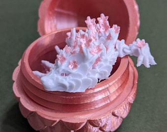 May include: A white, 3D printed dragon figurine with pink flowers on its back, inside a pink, 3D printed acorn-shaped box.
