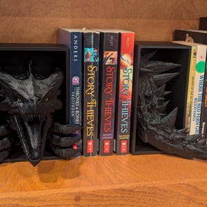 May include: Black dragon-themed bookends on a wooden shelf. One bookend features the dragon's head, the other its tail. Books with titles like "Story Thieves" and "Trickster's Queen" are visible between the bookends.