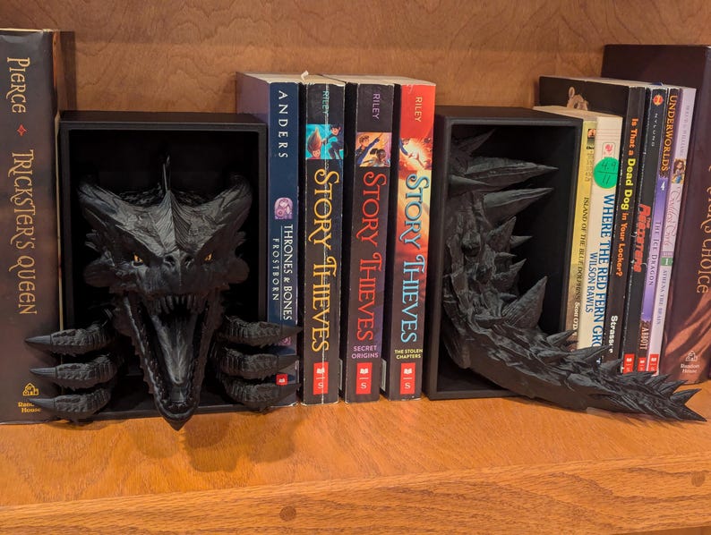 May include: Black dragon-themed bookends on a wooden shelf. One bookend features the dragon's head, the other its tail. Books with titles like "Story Thieves" and "Trickster's Queen" are visible between the bookends.