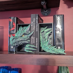 May include: Green dragon bookends with detailed scales and a fierce expression. The bookends are placed on a wooden shelf with books between them. The books include titles like "eclipse" and "breaking dawn".