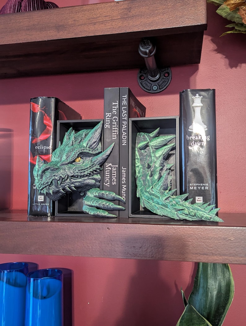 May include: Green dragon bookends with detailed scales and a fierce expression. The bookends are placed on a wooden shelf with books between them. The books include titles like "eclipse" and "breaking dawn".