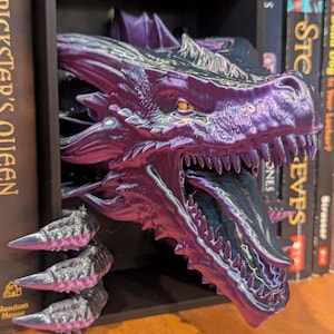 May include: A purple and black dragon head bookend with sharp teeth and a long tongue. The dragon is 3D printed and is emerging from a black shelf. Books are visible on either side of the bookend.