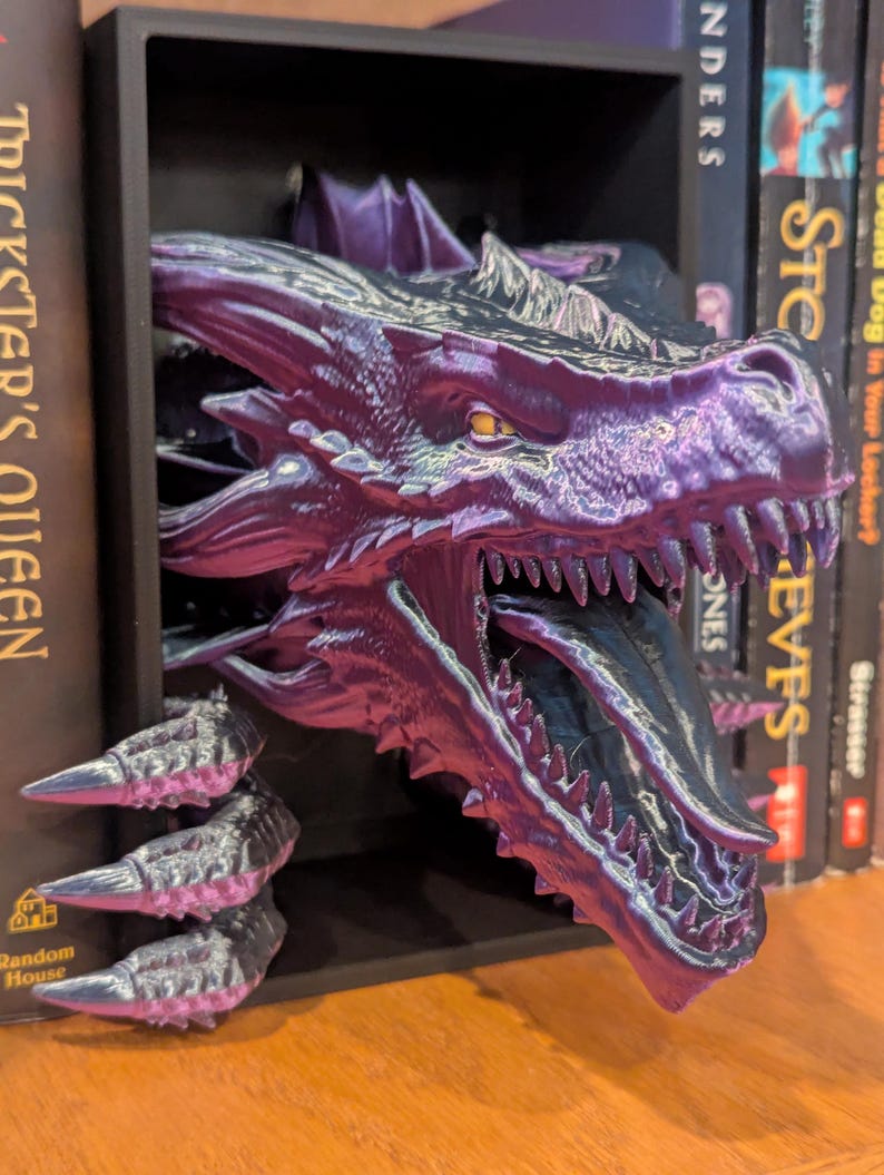May include: A purple and black dragon head bookend with sharp teeth and a long tongue. The dragon is 3D printed and is emerging from a black shelf. Books are visible on either side of the bookend.