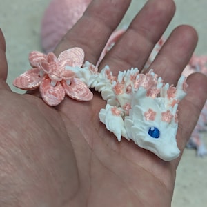 Puede incluir: Una peque&ntilde;a figura de drag&oacute;n blanco con detalles rosas y un ojo azul. El drag&oacute;n sostiene una flor rosa en su boca.