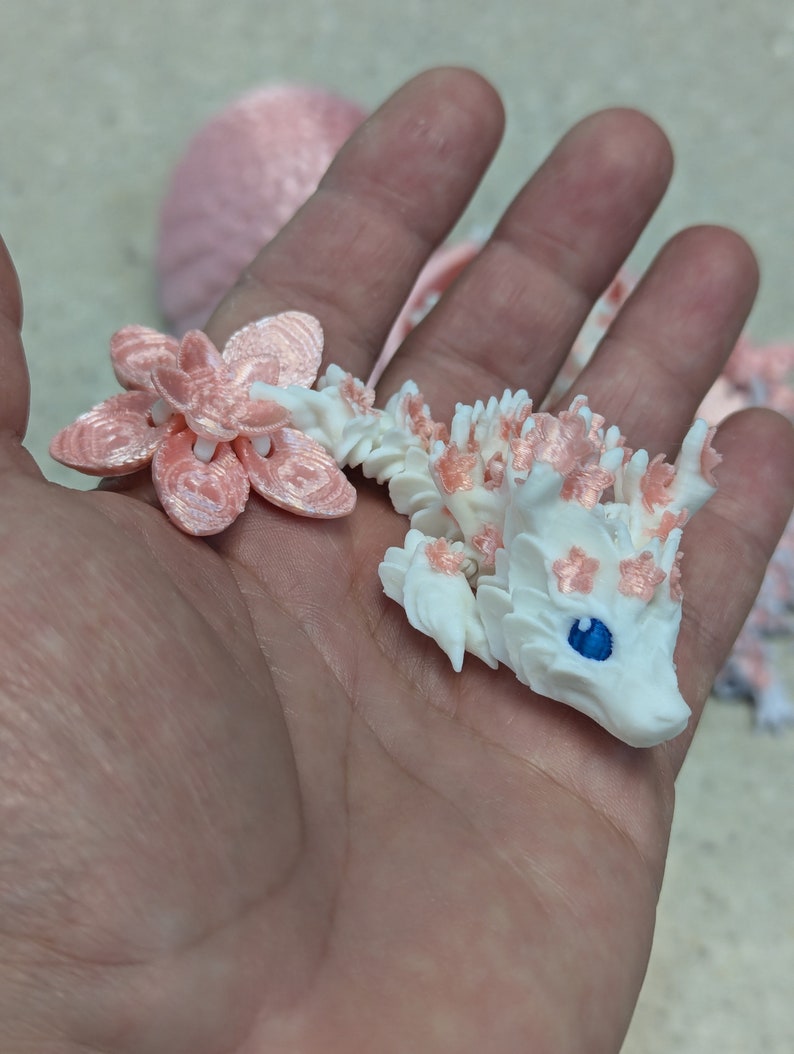 Puede incluir: Una peque&ntilde;a figura de drag&oacute;n blanco con detalles rosas y un ojo azul. El drag&oacute;n sostiene una flor rosa en su boca.