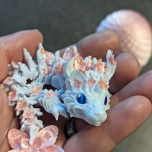 Puede incluir: Una figurita de drag&oacute;n blanca impresa en 3D con detalles de flores rosas. El drag&oacute;n tiene ojos azules y un cuerpo texturizado detallado.