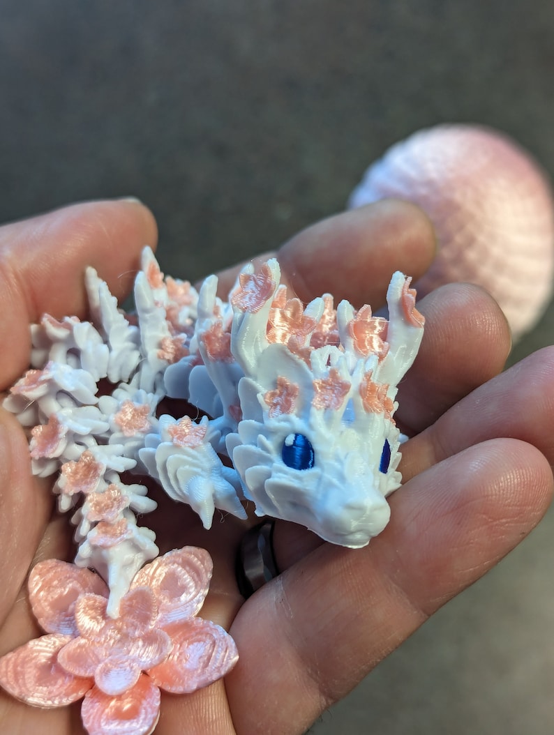 Puede incluir: Una figurita de drag&oacute;n blanca impresa en 3D con detalles de flores rosas. El drag&oacute;n tiene ojos azules y un cuerpo texturizado detallado.