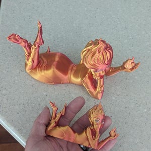 May include: Two orange and gold 3D-printed figurines of a reclining woman with flame-like hair and holding a flame. One figurine is held in a hand, the other is on a countertop.