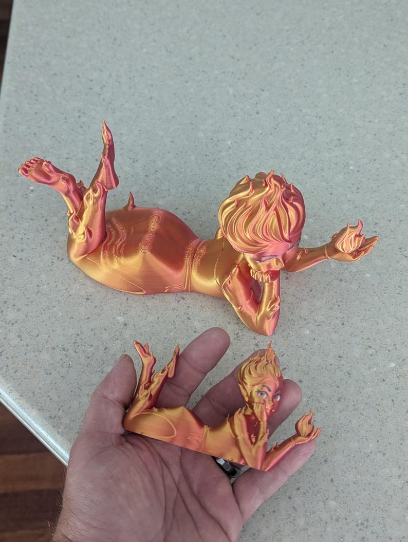 May include: Two orange and gold 3D-printed figurines of a reclining woman with flame-like hair and holding a flame. One figurine is held in a hand, the other is on a countertop.