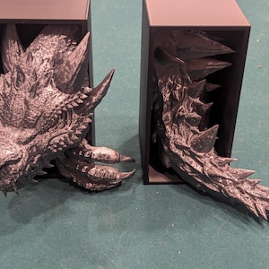 May include: Two gray dragon-themed bookends. One bookend features the dragon's head, with horns and detailed scales. The other bookend shows the dragon's tail, with sharp spikes. Both are set against a black background.