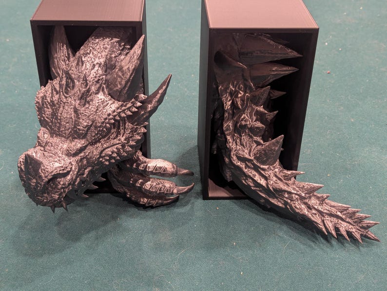 May include: Two gray dragon-themed bookends. One bookend features the dragon's head, with horns and detailed scales. The other bookend shows the dragon's tail, with sharp spikes. Both are set against a black background.