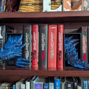 May include: A dark wooden bookshelf displays a collection of books, including "Breaking Dawn" and "The Vampire Diaries." Two blue dragon bookends flank the books. The books have red and black spines.
