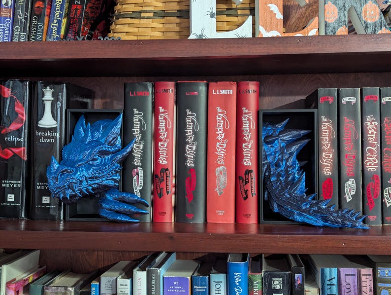 May include: A dark wooden bookshelf displays a collection of books, including "Breaking Dawn" and "The Vampire Diaries." Two blue dragon bookends flank the books. The books have red and black spines.