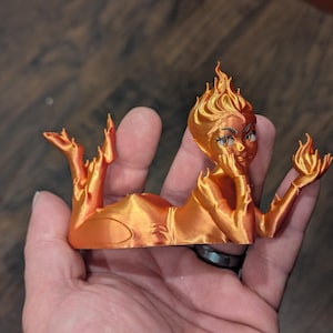 May include: A 3D-printed orange figurine of a reclining woman with flames for hair and holding a flame in her hand. The figurine has detailed facial features and is resting on a wooden surface.