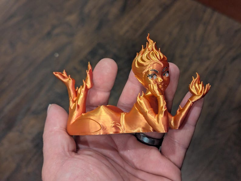 May include: A 3D-printed orange figurine of a reclining woman with flames for hair and holding a flame in her hand. The figurine has detailed facial features and is resting on a wooden surface.