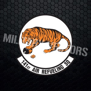 May include: A white circle with a black outline and the text "141st Air Refueling Sq" around the edge. Inside the circle is an orange tiger with black stripes, pouncing on a black object.