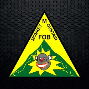 Pode incluir: Um triângulo amarelo com uma montanha verde e um macaco de desenho animado com olhos vermelhos e um grande sorriso. O texto "Monkey Mountain FOB" está escrito em torno do triângulo.