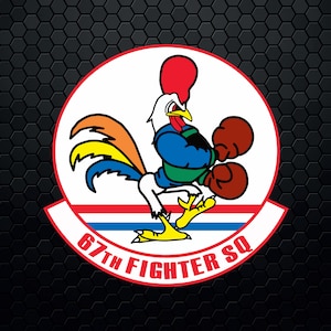 May include: A red, white, and blue emblem with a rooster wearing boxing gloves. The rooster is facing left and has a yellow, orange, and red tail. The text "67th Fighter SQ" is written in a red, white, and blue arc below the rooster.