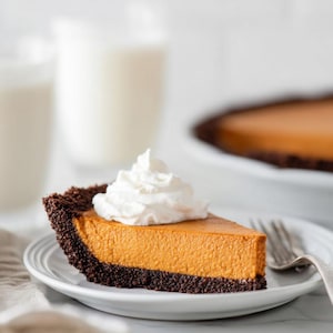 Puede incluir: Una porción de tarta de calabaza con crema batida en un plato blanco. La tarta tiene una corteza marrón oscuro y un relleno de color naranja brillante. Un tenedor descansa en el plato y una tarta entera está en el fondo. Dos vasos de leche están borrosos en el fondo.