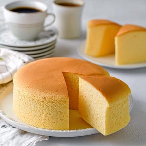 Op de afbeelding: Een goudbruine, ronde cake met een uitgesneden stuk, gepresenteerd op een witte bord. Twee extra plakken liggen op een apart bord. Twee kopjes koffie en schotels op de achtergrond, wat duidt op een dessert.
