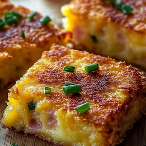 May include: Close-up of golden-brown, square-shaped food items, possibly appetizers or snacks. The food has a crispy exterior and a soft, yellow interior with visible ham. Garnished with fresh green chives, the dish is presented on a wooden surface.