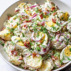 May include: A bowl of potato salad featuring boiled eggs, red onion, bacon pieces, and fresh dill. The creamy dressing coats the potatoes and other ingredients, creating a colourful and appetising dish. The salad is served in a white bowl.
