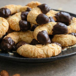 May include: A plate of crescent-shaped biscuits, coated with flaked almonds and dipped in dark chocolate. The biscuits are arranged on a dark plate, with a few almonds scattered around. A delicious treat.