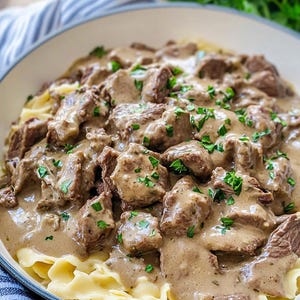 Peut inclure: Un bol de bœuf stroganoff composé de morceaux de bœuf bruns et tendres dans une sauce crémeuse brun clair, servis sur des nouilles aux œufs jaunes. Le plat est garni de persil vert frais et présenté dans un bol blanc large et peu profond.
