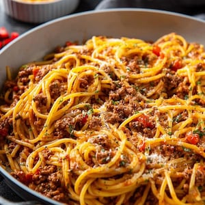 Puede incluir: Primer plano de espaguetis con salsa de carne en una sartén. La pasta es de color amarillo dorado, y la salsa es de color marrón oscuro con trozos de tomate. Un pequeño cuenco de queso rallado está en el fondo.