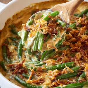 May include: A close-up of a casserole dish filled with green beans in a creamy sauce, topped with crispy fried onions. A wooden spoon scoops up a portion of the dish, showcasing the textures and colors of the food. The dish is in a white ceramic baking dish.