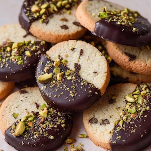 May include: A stack of shortbread biscuits, partially coated in dark chocolate and topped with chopped pistachios. The circular biscuits are light brown with a textured surface. Some have chocolate chips. The image has a warm colour palette.