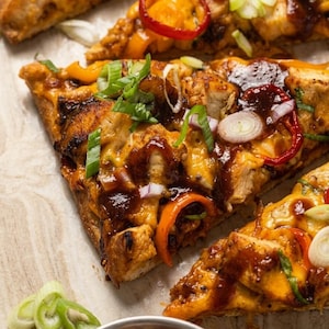May include: Close-up of several slices of pizza with toppings. The pizza has a dark crust, melted cheese, chicken, red and orange bell peppers, red onion, and green onions. A small bowl of red pepper flakes is in the foreground.
