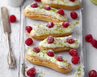 Könnte beinhalten: Draufsicht auf mehrere Eclairs auf einem silbernen Tablett. Jeder Eclair ist mit weißem Zuckerguss, frischen Himbeeren und Limettenschale belegt. Eine kleine Schüssel mit Himbeeren und ein Messer sind ebenfalls vorhanden, zusammen mit verstreuten Himbeeren.