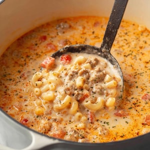 May include: A close-up shot of a creamy soup with elbow macaroni, ground meat, and diced tomatoes. A black ladle scoops up a portion of the soup from a white pot. The soup has a light orange hue and is garnished with herbs.