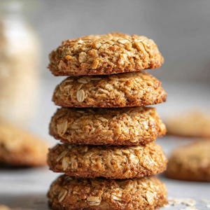 Puede incluir: Una pila de cuatro galletas de avena de color marrón dorado. Las galletas son redondas y tienen una superficie texturizada con avena visible. Las galletas están apiladas una encima de la otra, con algunas avenas sueltas esparcidas en la base. El fondo es de color neutro.