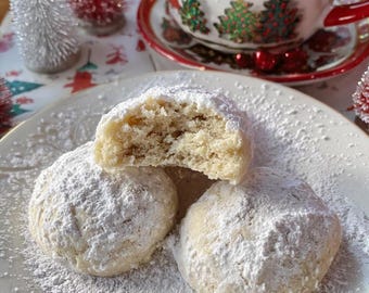 Könnte beinhalten: Drei mit Puderzucker bestäubte Shortbread-Kekse auf einem weißen Teller, einer davon ist zerbrochen. Im Hintergrund eine festliche rot-weiß-Tasse und Untertasse mit Weihnachtsschmuck.