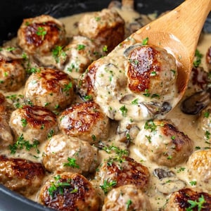Peut inclure: Gros plan sur des boulettes de viande dans une sauce crémeuse aux champignons, garnies d'herbes fraîches, dans une poêle noire. Une cuillère en bois ramasse plusieurs boulettes. Les boulettes sont dorées et semblent juteuses.