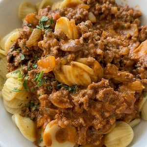 Könnte beinhalten: Nahaufnahme einer weißen Schüssel mit Pasta und Fleischsoße. Die Pasta hat eine Muschelform, und die Soße ist von einer satten braunen Farbe mit sichtbaren Fleisch- und Gemüsestücken. Frische grüne Kräuter sind darüber gestreut.