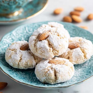 Peut inclure: Gros plan sur des biscuits aux amandes saupoudrés de sucre glace, chacun garni d'une amande entière, disposés sur une assiette turquoise. Une tasse et une soucoupe assorties sont en arrière-plan, avec des amandes éparpillées.