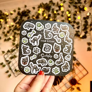 May include: A black and white sticker sheet featuring illustrations of black cats in various poses. The sheet has a black background with white outlines and includes the text "void kitties" and "Squidly Ink".