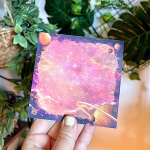 Può includere: Blocco note quadrato con un design a tema spaziale. La nota presenta uno sfondo galattico rosa e viola con pianeti e stelle. I bordi sono blu scuro e l'estetica generale è stravagante e artistica.